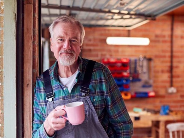A man in a garage holding a cup of tea and wearing overalls.